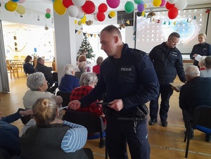 dwaj policjanci dzielnicowi rozdają siedzącym na krzesłach seniorom opaski odblaskowe