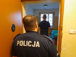 w celi pomieszczenia dla osób zatrzymanych stoi mężczyzna. Za nim przy otwartych drzwiach stoi policjant służby dyżurnej