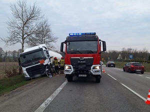 miejsce zdarzenia drogowego. W rowie znajduje się przechylony TIR z cysterną. Obok na jezdni stoi wóz straży pożarnej. Przy tirze stoją strażacy