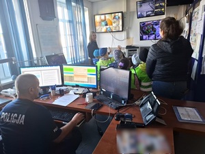 grupa przedszkolaków w pomieszczeniu dyżurnego komendy lęborskiej policji przyglądająca się wiszącym na ścianie monitorom. Przed dziećmi stoi policjantka, za dziećmi na stanowisku kierowania siedzi dyżurny