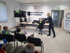 w auli budynku komendy lęborskiej policji policjantka nakłada kask ochronny przedszkolakowi ubranemu w sprzęt przeciwuderzeniowy