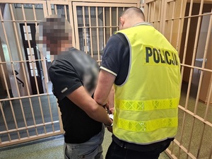 w okratowanym pomieszczeniu dla osób zatrzymanych policjanta zakłada na ręce mężczyzny kajdanki