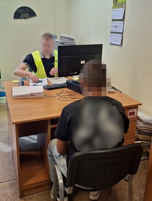 policjant przesłuchuje siedzącego na krześle obok biurka mężczyznę