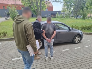 przed nieoznakowanym radiowozem stoją dwaj nieumundurowani policjanci i mężczyzna, który ma ręce skute za plecami kajdankami. Policjant otwiera drzwi radiowozu