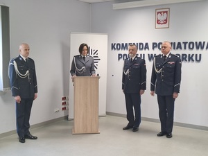 w auli lęborskiej komendy policji stoją przy ścianie z napisem Komenda Powiatowa Policji w Lęborku Komendant Powiatowy Policji w Lęborku oraz Komendant Wojewódzki Policji w Gdańsku. Obok przy mównicy stoi policjantka, obok niej policjant - dowódca uroczystości