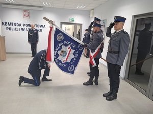 w auli lęborskiej komendy policji Komendant Powiatowy Policji w Lęborku klęka przy sztandarze trzymanym przez policjantów pocztu sztandarowego, którzy salutują