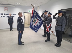 w auli lęborskiej komendy policji Komendant Powiatowy Policji w Lęborku stoi przed sztandarem trzymanym przez policjantów pocztu sztandarowego, którzy salutują