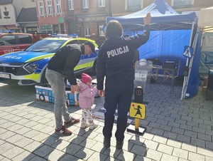 na placu przed policyjnym stoiskiem i przed zabawkowym przejściu dla pieszych stoi policjantka z uniesioną prawą ręką w górę i trzymająca za rękę małą dziewczynkę. Obok dziewczynki stoi jej ojciec