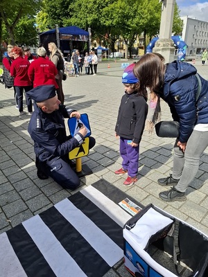 na placu  przy zabawkowym przejściu dla pieszych policjant objaśnia znaczenie zabawkowego znaku drogowego małemu chłopcu