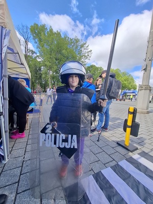 na placu przed policyjnym stoiskiem stoi chłopiec ubrany w sprzęt ochronny wykorzystywany przez policjantów podczas zabezpieczania imprez masowych
