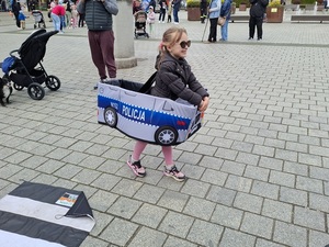 placem idzie dziewczynka w autochodziku  zawieszonym na jej ramionach
