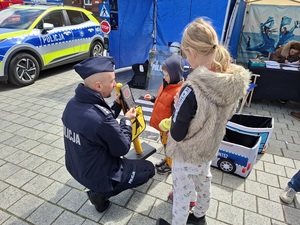 na placu przed policyjnym stoiskiem przy zabawkowym przejściu dla pieszych klęczy policjant który objaśnia chłopcu znacznie zabawkowego znaku drogowego