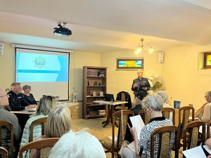 debata w sali biblioteki miejskiej. Przed siedzącymi na krzesłach seniorami stoi policjantka, moderatorka spotkania. Obok przy stole siedzi zastępca komendanta powiatowego policji w Lęborku, dzielnicowy i przedstawicielka lęborskiego starostwa. Na ekranie zawieszonym na ścianie wyświetla się prezentacja