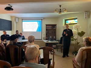 debata w sali biblioteki miejskiej. Przed siedzącymi na krzesłach seniorami stoi policjantka, moderatorka spotkania. Obok przy stole siedzi zastępca komendanta powiatowego policji w Lęborku, dzielnicowy i przedstawicielka lęborskiego starostwa. Na ekranie zawieszonym na ścianie wyświetla się prezentacja