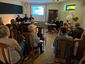 debata w sali biblioteki miejskiej. Przed siedzącymi na krzesłach seniorami siedzi przedstawicielka Starostwa Powiatowego w Lęborku. Obok przy stole siedzi zastępca komendanta powiatowego policji w Lęborku, dzielnicowy i policjantka - moderatorka spotkania. Na ekranie zawieszonym na ścianie wyświetla się prezentacja.