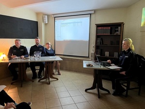 debata w sali biblioteki miejskiej. Przed siedzącymi na krzesłach seniorami siedzi przy laptopie policjantka, moderatorka spotkania. Obok przy stole siedzi zastępca komendanta powiatowego policji w Lęborku, dzielnicowy i przedstawicielka lęborskiego starostwa. Na ekranie zawieszonym na ścianie wyświetla się prezentacja. Komendant zabiera głos