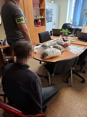 w policyjnym pomieszczeniu służbowym na krześle siedzi zatrzymany mężczyzna. Przed nim na stole leżą opakowania foliowe wypełnione suszem roślinnym. Obok stoi nieumundurowany policjant z opaską na ramieniu z napisem policja