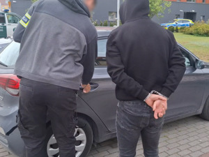 policjant kryminalny otwiera tylne drzwi nieoznakowanego radiowozu. Obok stoi mężczyzna, który ma ręce skute za plecami kajdankami