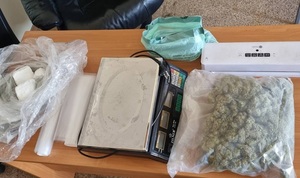 na stole leży białe foliowe opakowanie z trzema grudami białego proszku. Obok waga elektroniczna, zgrzewarka, rolka folii i dwa duże opakowania z zielonym suszem roślinnym