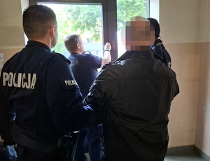 dwoje policjantów służby konwojowej wyprowadza z budynku komendy zatrzymanego mężczyznę. Drzwi otwiera policjant dyżurny