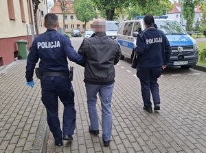 policjant służby konwojowej prowadzi w kierunku radiowozu zatrzymanego mężczyznę. Obok idzie policjantka