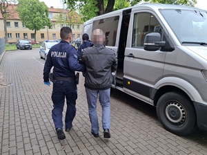policjant służby konwojowej prowadzi w kierunku uchylonych bocznych drzwi nieoznakowanego radiowozu zatrzymanego mężczyznę