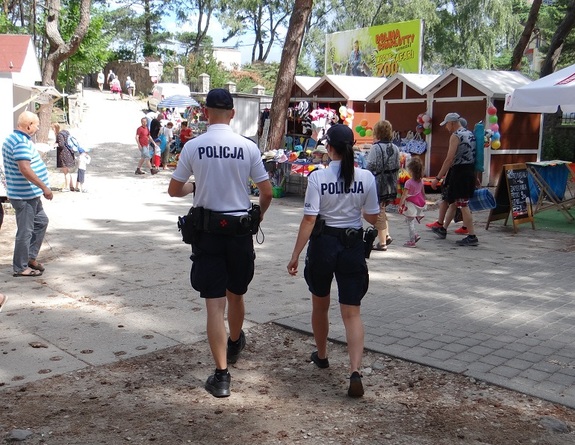 policjantka i policjant w letnim umundurowaniu (czarne spodenki, białe koszulki polo) idący pośród turystów w stronę wejścia na plażę nad morzem