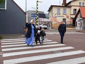 grupa przedszkolaków i siostra zakonna, która trzyma przedszkolaka za rękę i podnosi drugą do góry przechodzą przez przejście dla pieszych. Obok idzie policjantka