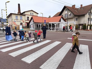 przez przejście dla pieszych przechodzą przedszkolaki z rękoma uniesionymi w górę. Jednego przedszkolaka trzyma za rękę siostra zakonna. Obok przejścia stoi policjantka, która obserwuje dzieci