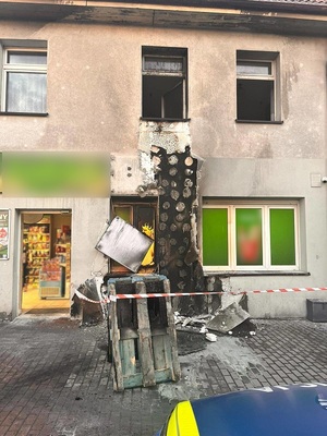 Budynek po pożarze. Na chodniku przy ścianie leżą drewniane nadpalone palety ogrodzone taśmą. Na ich wysokości spalony jest pionowy fragment elewacji. Ślady pożaru prowadzą do dwóch okien. Przed budynkiem stoi policyjny radiowóz