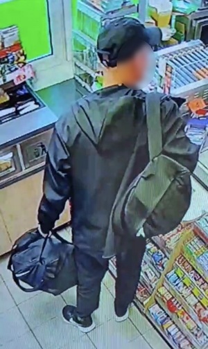 mężczyzna w czarnej kurtce, czarnych spodniach i czarnej czapce z daszkiem przed ladą sklepową. Trzyma torbę podróżną, na plecach ma plecak