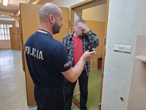 w korytarzu pomieszczenia dla osób zatrzymanych przy otwartych drzwiach celi stoi mężczyzna. Obok stoi policjant dyżurny, który pokazuje mężczyźnie wynik badania alkotestem