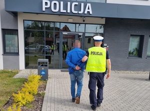 policjant ruchu drogowego prowadzi w kierunku wejścia do budynku komisariatu policji mężczyznę, który ma ręce skute za plecami kajdankami
