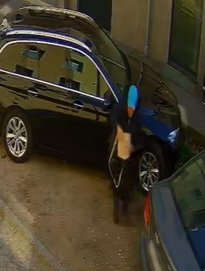 kobieta na ulicy obok czarnego suva, ubrana w czarną kurtkę, ciemne spodnie i niebieską czapkę z daszkiem, trzymająca kremową torebkę