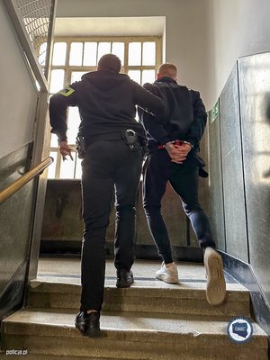 policjant prowadzący schodami w górę mężczyznę, który ma ręce skute za plecami kajdankami