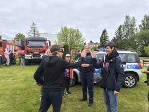 festyn. Na tle radiowozu przy którym stoi policjantka nastolatek w narkogoglach przybija piątkę koledze