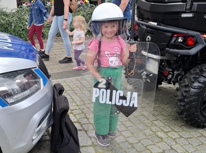 festyn. Obok radiowozu stoi kilkuletnia dziewczynka z policyjną tarczą, pałką i kaskiem na głowie