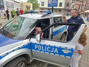 festyn. W radiowozie na miejscu kierowcy siedzi uśmiechnięta dziewczynka. Przed nią stoi policjant