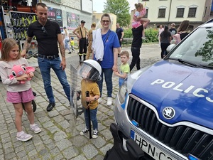 festyn. Przy radiowozie stoi mały chłopiec z policyjną tarczą, pałką i z kaskiem na głowie. Obok stoją jego rodzice i siostra