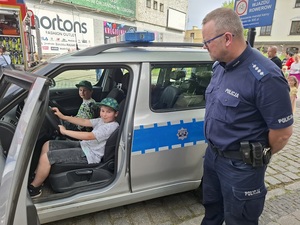 w radiowozie na miejscu kierowcy i pasażera siedzą dwaj chłopcy. Obok stoi policjant
