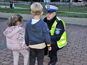 na chodniku w pobliżu placu zabaw policjant ruchu drogowego kuca przed kilkuletnią dziewczynką i kilkuletnim chłopcem
