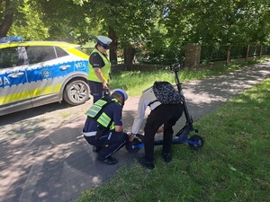 na drodze dla rowerów policjant ruchu drogowego sprawdza stan techniczny hulajnogi elektrycznej, przy której stoi młody mężczyzna. Obok stoi policjantka