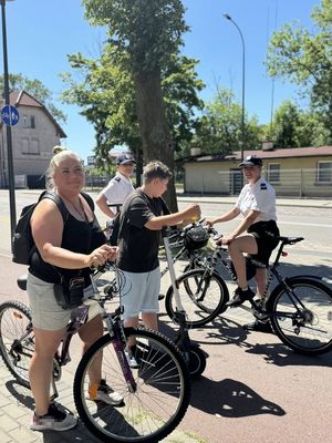 dwoje uśmiechniętych policjantów na rowerach obok chłopca na hulajnodze elektrycznej i kobiety na rowerze