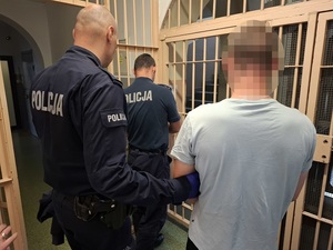 dyżurny policji otwiera zakratowane drzwi. Za nim stoi mężczyzna i policjant, który przytrzymuje go za ramię