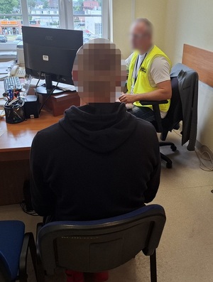 w pokoju przesłuchań na krześle siedzi zatrzymany mężczyzna. Przed nim przy biurku i przed komputerem siedzi policjant