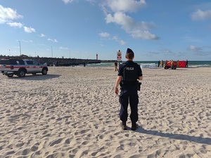 na nadmorskiej plaży stoi policjant. W tle widać czerwony parawan ratowników wodnych, SAR, strażaków. Na plaży stoi radiowóz straży pożarnej