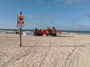 na nadmorskiej plaży w miejscu ustawienia znaku zakaz kąpieli, przy brzegu znajduje się czerwony parawan przy którym stoją ratownicy wodni, SAR i strażacy. Przy parawanie zaparkowany jest samochód SAR i quad ratowników wodnych