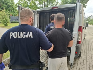 z tyłu nieoznakowanego radiowozu policjant trzyma za ramię zatrzymanego mężczyznę. Drugi policjant otwiera kratę w drzwiach radiowozu
