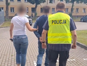 na zewnątrz budynku komendy dwoje policjantów prowadzi w kierunku radiowozów mężczyznę, który ma ręce skute za plecami kajdankami