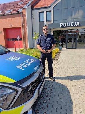 przed budynkiem komisariatu Policji w Łebie i obok radiowozu stoi umundurowany policjant w stopniu młodszego aspiranta